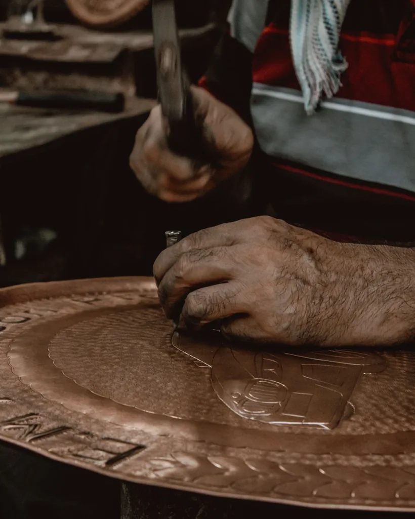 Detalle de un cincel apoyado sobre una chapa metálica mientras el martillo repasa el trazo del relieve.