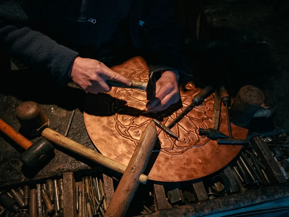 Manos de un artesano cincelando el relieve de una bandeja metálica apoyada sobre una almohadilla, con martillo y cinceles sobre la mesa del taller.